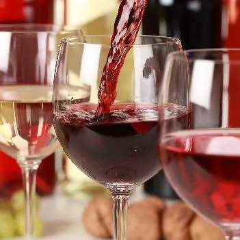 Auf dem Bild sind drei Gläser Wein zu sehen. In das mittlere Glas wird ein Rotwein eingeschenkt. In den anderen beiden Gläsern befinden sich Rotwein und Weißwein. Im Hintergrund sind gefüllte Weinflaschen zu erkennen.
